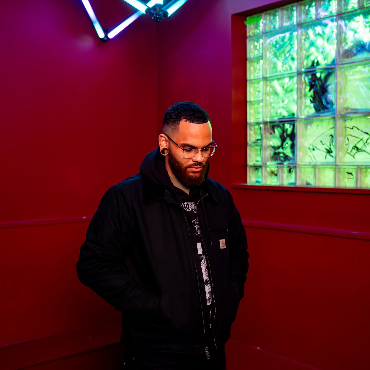 Manwell in a black jacket and glasses standing in a red room by a glass block window
